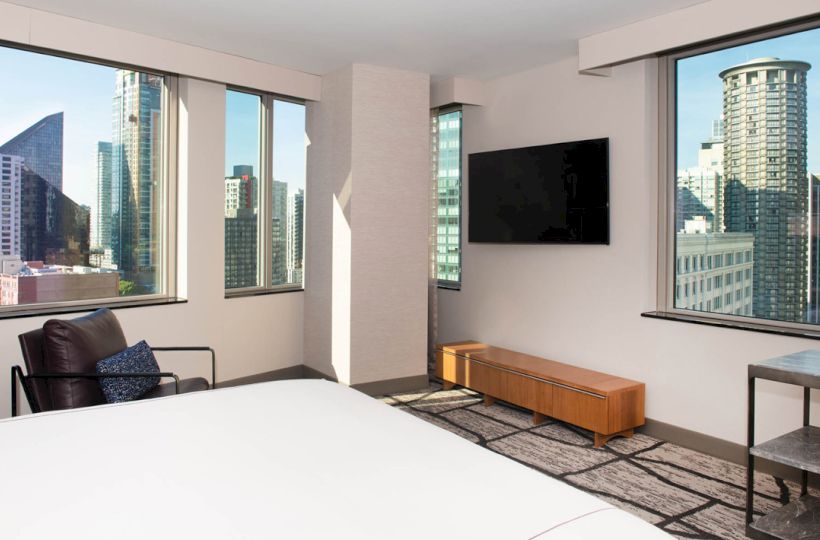 A modern hotel room with large windows showing a city view, featuring a bed, chair, TV, and minimalist decor.