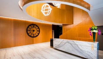 A modern lobby features a curved wooden staircase, large clock, marble reception desk, and decorative lights and flowers.