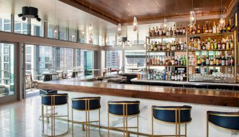 The image shows a modern bar with high stools and a fully stocked liquor shelf, featuring large windows and a wooden ceiling.