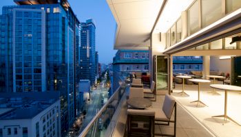 A modern urban balcony at dusk, featuring seating and tables with a cityscape view and contemporary lighting.