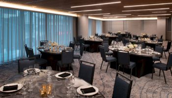 The image shows an elegant dining area with round tables set for a formal event, featuring chairs, plates, wine glasses, and floral centerpieces.