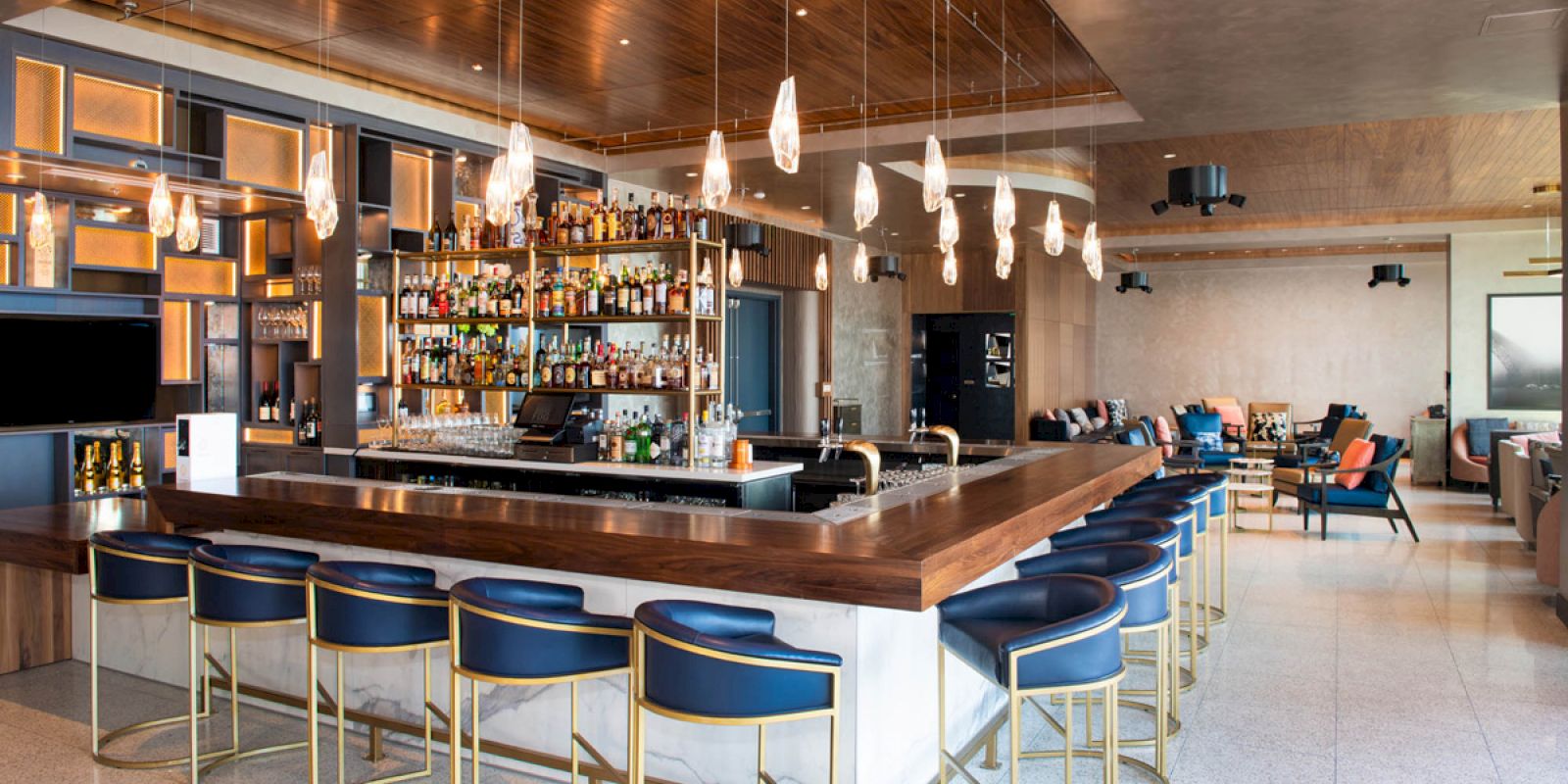 This image shows a modern bar with blue stools, hanging lights, and a well-stocked counter, in an elegant and spacious seating area.