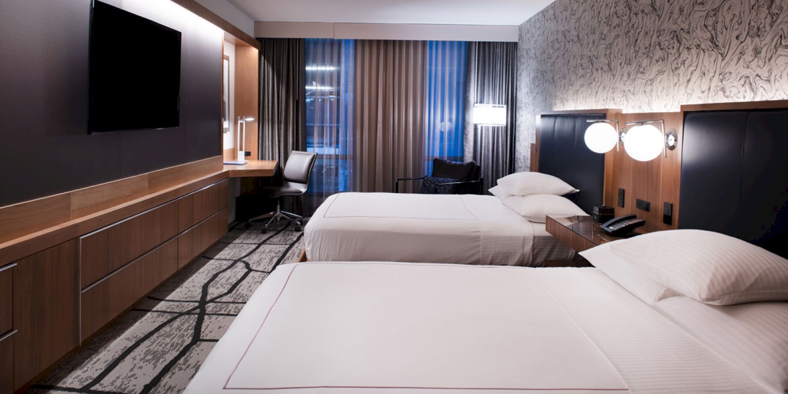 A modern hotel room with two beds, a wall-mounted TV, desk with chair, and patterned carpet, featuring stylish lighting and decor.