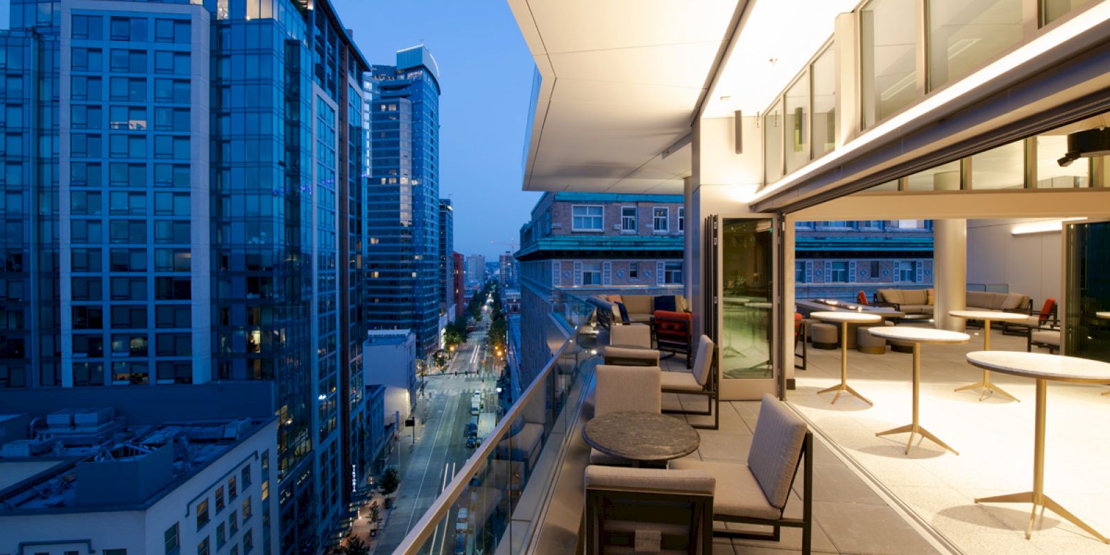 A modern rooftop terrace with seating overlooks a city street lined with tall buildings at dusk, offering an urban view.
