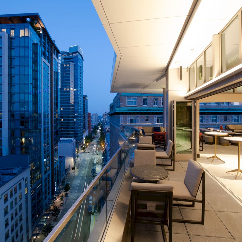 A modern rooftop terrace with seating, overlooking a cityscape with tall buildings at dusk. The area is well-lit, creating a cozy atmosphere.