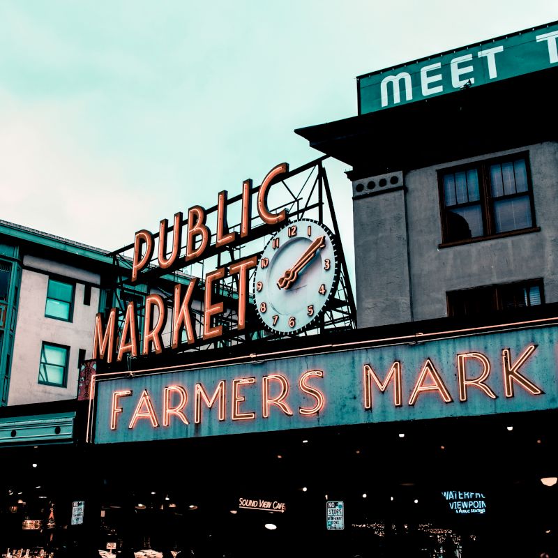 The image shows a neon sign for a "Public Market" and "Farmers Market" alongside a clock, set on an urban building backdrop.