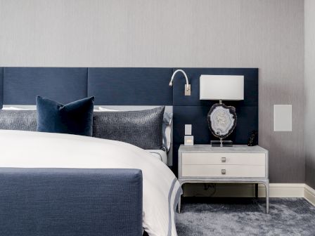 A stylish bedroom with a navy blue headboard, white bedding, gray pillows, and a nightstand with a lamp and decorative items.