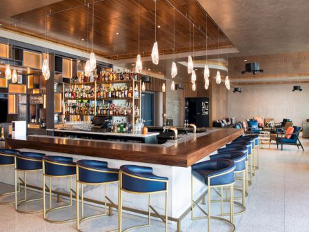 The image shows a modern bar with blue stools, hanging lights, and a well-stocked bar counter in a spacious, stylish lounge area.