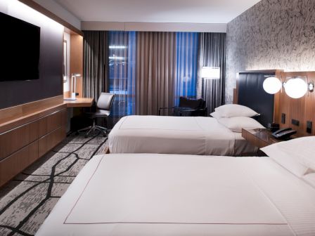 The image shows a modern hotel room with two beds, a wall-mounted TV, a desk with a chair, and stylish decor.