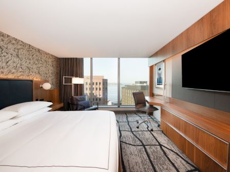 The image shows a modern hotel room with a large bed, a desk, a chair, and a big wall-mounted TV. There's a window with city views.