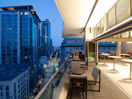 A modern outdoor terrace overlooking a cityscape at dusk, featuring seating and tables, adjacent to a lit indoor space.