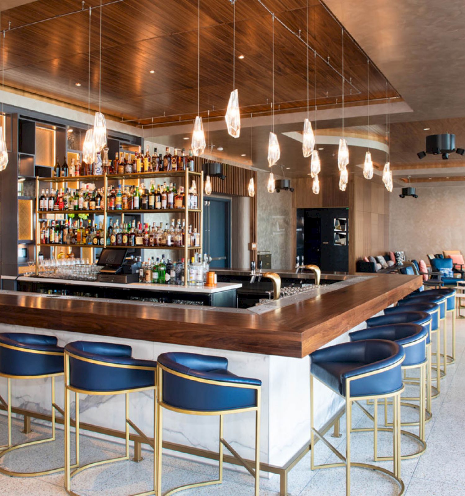 The image shows a modern bar with a wooden counter, blue chairs, and hanging lights, set in a stylish lounge area.
