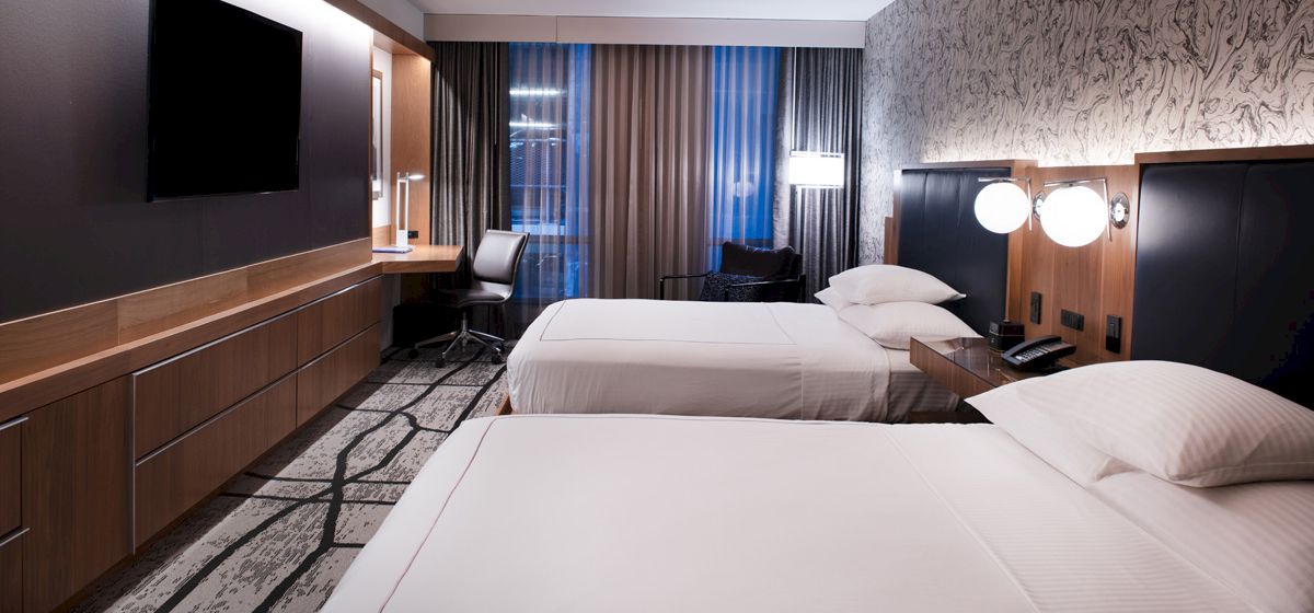 A modern hotel room with two double beds, a flat-screen TV, desk, and a window with curtains. The lighting is warm and inviting.