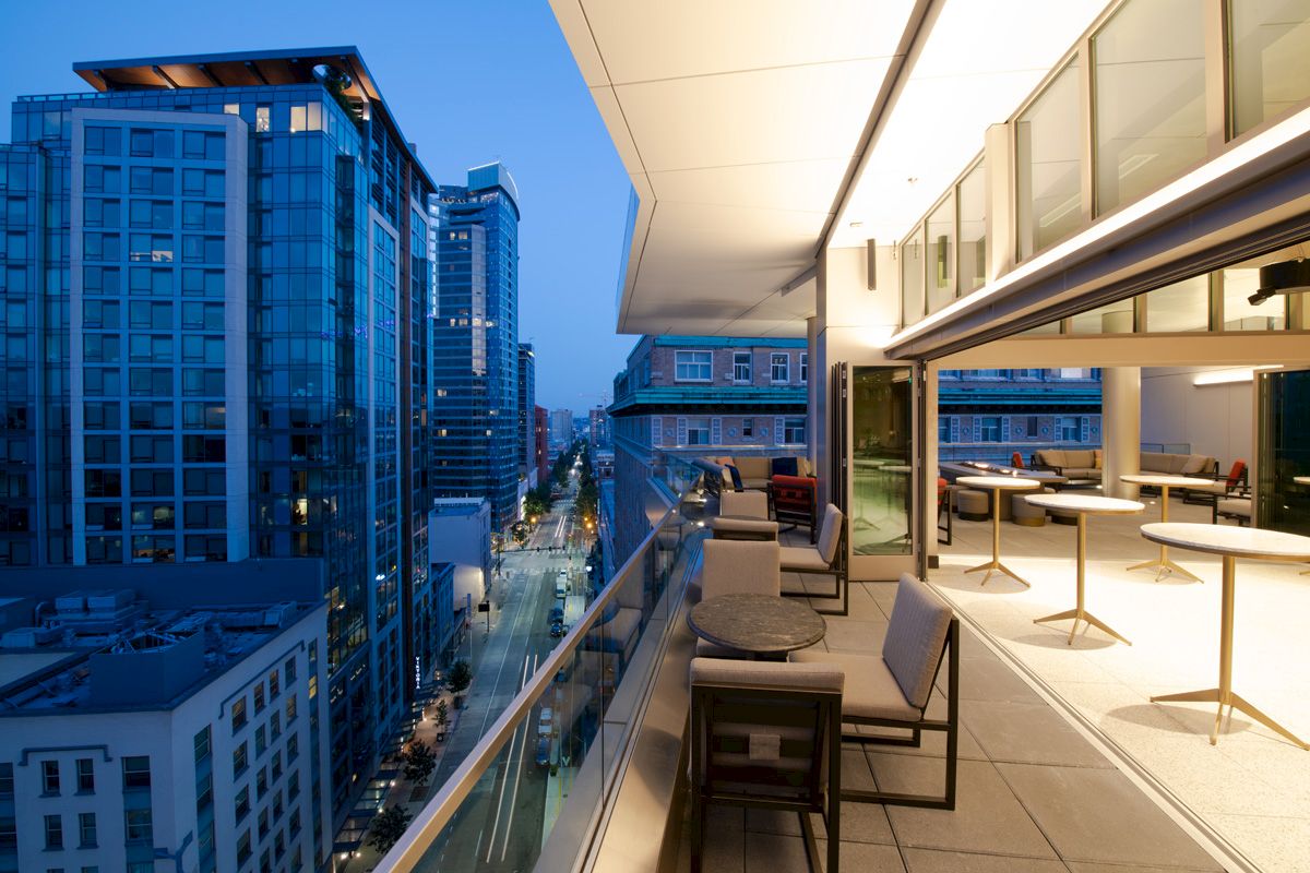 The image shows a modern urban balcony view at dusk with seating, overlooking a cityscape with high-rise buildings and illuminated streets.