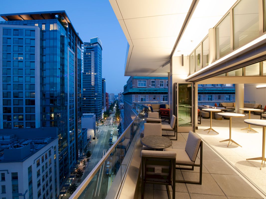 The image shows a modern urban balcony view at dusk with seating, overlooking a cityscape with high-rise buildings and illuminated streets.