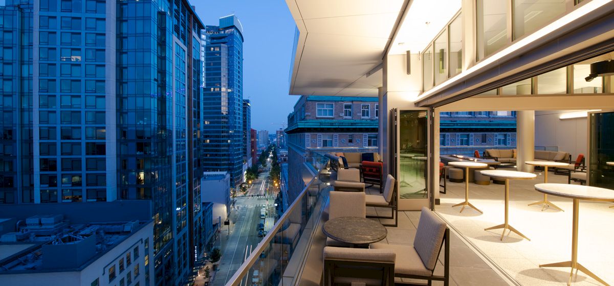 The image shows a modern urban balcony view at dusk with seating, overlooking a cityscape with high-rise buildings and illuminated streets.