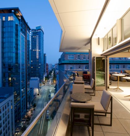 A modern rooftop terrace overlooks a city street, featuring seating areas with tables, surrounded by tall buildings in the evening light.
