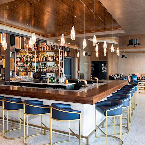 This image shows a modern bar with stylish seating, a well-stocked bar, hanging lights, and a lounge area in the background.