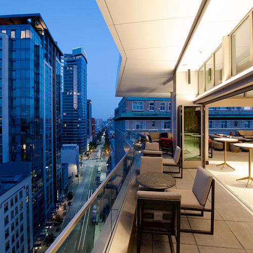 A cityscape at dusk with a modern balcony featuring tables and chairs, high-rise buildings lining the streets, and soft ambient lighting.