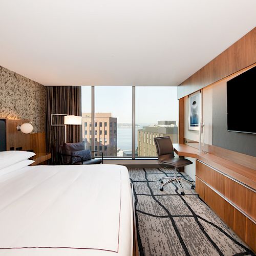 A modern hotel room features a large bed, a desk, a flat-screen TV, and a city view through a wide window.