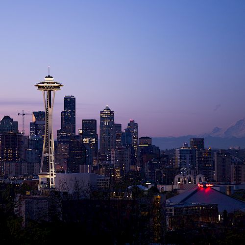 A city skyline at dusk is shown, featuring a tall tower and mountains in the distance, with buildings illuminated against a fading sky.