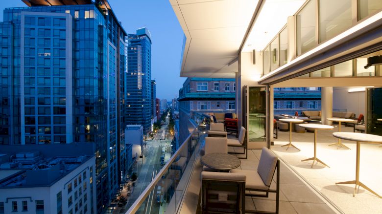 A rooftop terrace with seating overlooks a city street lined with modern buildings at dusk. The scene is well-lit and inviting.