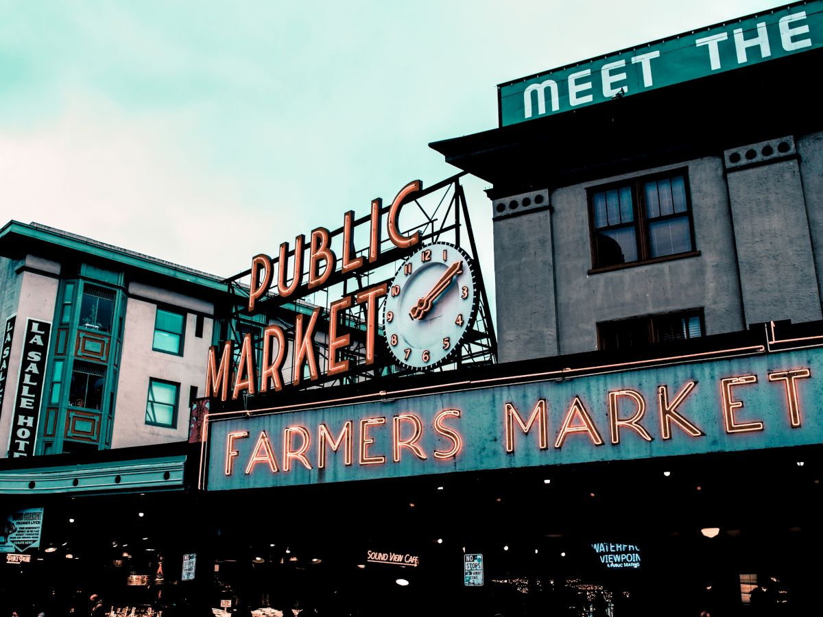 The image shows the iconic neon signs of a "Public Market" and "Farmers Market" with a clock, against the backdrop of surrounding buildings.