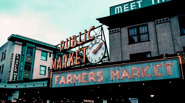 The image shows the iconic neon signs of a "Public Market" and "Farmers Market" with a clock, against the backdrop of surrounding buildings.
