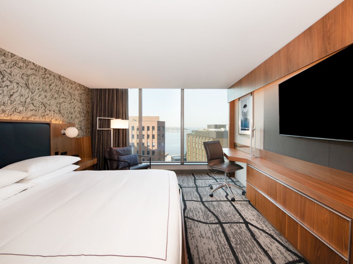 A modern hotel room with a large bed, desk, chair, TV, and a view of buildings through a window.