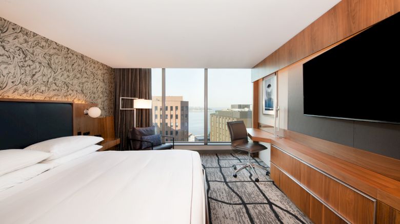 A modern hotel room with a large bed, desk, chair, TV, and a view of buildings through a window.