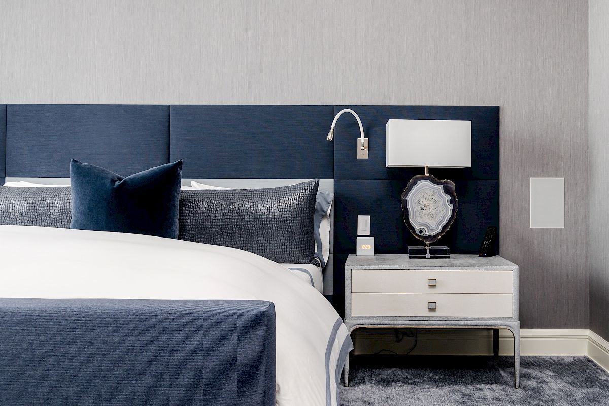 A modern bedroom with a blue and gray theme features a bed, side table with a lamp, and textured carpet below.
