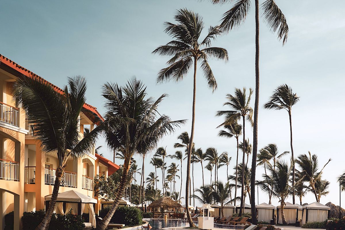 A tropical poolside scene featuring tall palm trees, lounge chairs, and a building with a red-tiled roof under a clear blue sky.