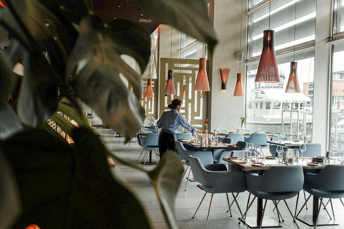 A person is setting tables in a modern restaurant with large windows, stylish lighting, and plants in the foreground.
