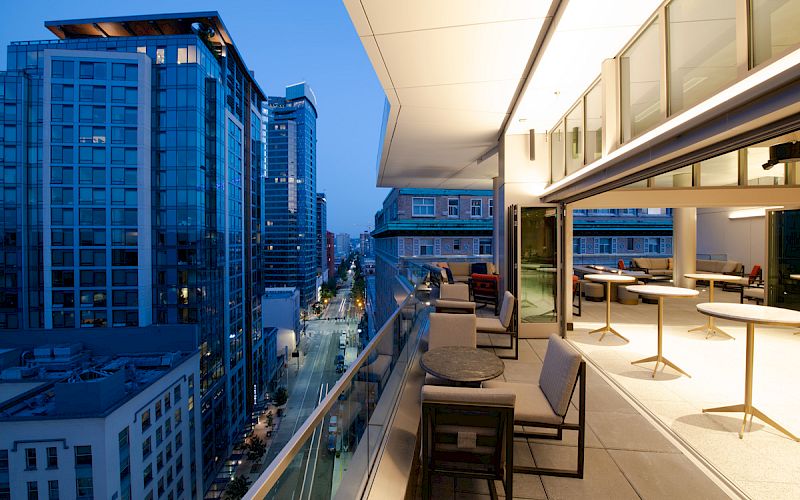 The image shows a modern rooftop patio with seating, overlooking an evening cityscape of tall buildings and illuminated streets.
