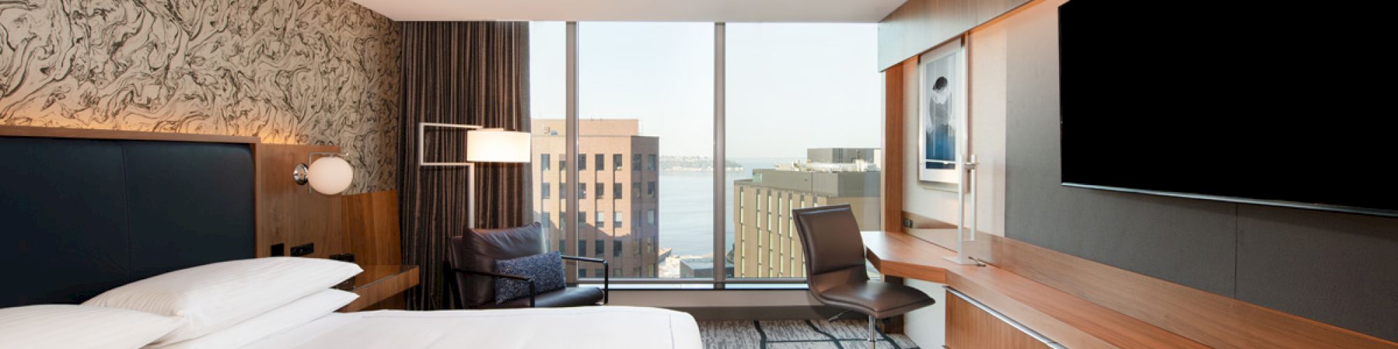 A modern hotel room with a large bed, flat-screen TV, desk, and chair. Large window offers a cityscape view.